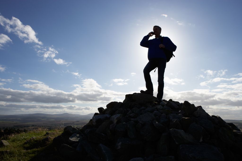Stock image of a man using a mobile phone atop a hill/mountain in a rural setting. Image credit: Shutterstock.