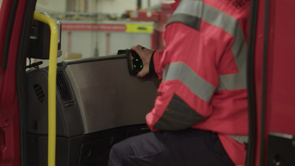 Body worn camera shown in front of a fire appliance held by firefighter shown from the side.