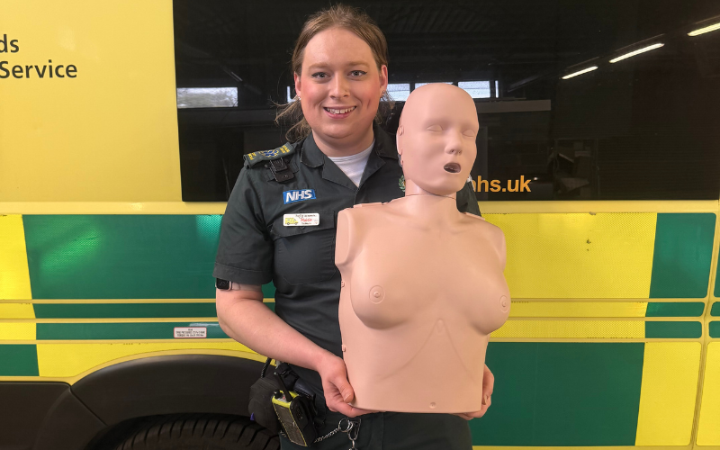 Maisie Stimson holds a female form mannequin in front of an EMAS ambulance.